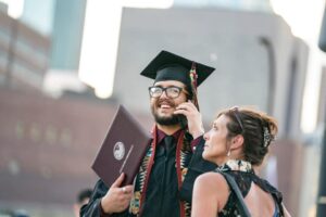 Student Joe Gaskill dressed in graduated robes holds his diploma while talking on phone